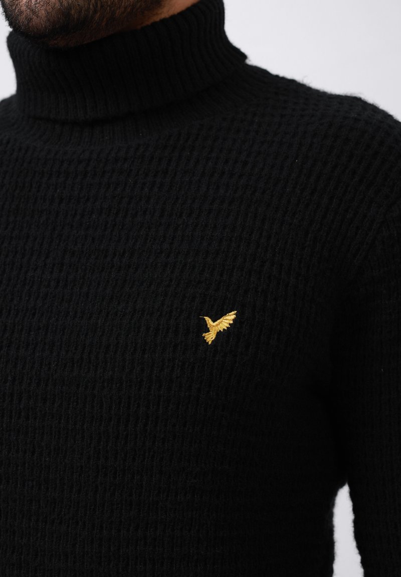 Maglione nero a collo alto lavorato a maglia con un motivo testurizzato e un piccolo logo dorato a forma di uccello sul lato sinistro del petto.