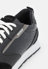 Sneaker noire avec un design lisse et texturé, comportant un côté gris à motif, des lacets blancs et une semelle blanche. Ornée du logo "HUGO".