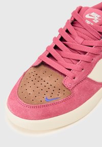 Nike SB sneaker, rózsaszín velúrból készült felsőrésszel, barna perforált orral és egy kis kék logóval. Lapos rózsaszín fűzőkkel és krém színű gumitalppal.