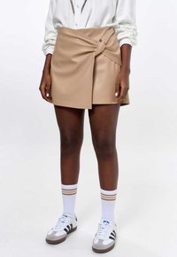 Beige neplederen wikkelrok met een geknoopt ontwerp en een drukknoopsluiting. Geselecteerd met witte sokken en grijze sneakers met zwarte strepen.