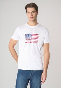 T-shirt bianca in cotone con una stampa logora della bandiera americana in rosso, bianco e blu, con maniche corte e collo rotondo.