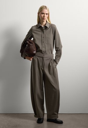 Una donna con lunghi capelli biondi indossa una camicia grigia sbottonata e pantaloni a gamba larga, mentre tiene una grande borsa di pelle marrone, in piedi contro un muro bianco.