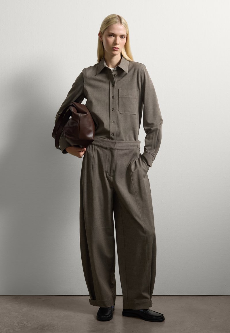Vrouw met lang blond haar draagt een losse grijze blouse met knopen en wijde broek, houdt een grote bruine leren tas vast en staat tegen een witte muur.