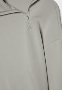 Haut à manches longues gris clair avec une courte fermeture éclair sur la poitrine et un détail de couture sur le haut du bras.
