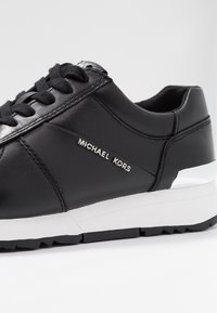 Zwarte leren sneaker met een gladde afwerking, voorzien van een witte zool, zilveren "Michael Kors"-logo en een gevoerde kraag voor comfort.