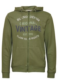 Sudadera con cremallera en verde oliva, con capucha ajustable, bolsillos frontales y texto impreso en navy y blanco. Textura suave de mezcla de algodón.
