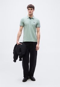 Νεαρός άνδρας που φοράει ανοιχτό πράσινο πόλο μπλουζάκι, μαύρο παντελόνι και μαύρα παπούτσια, κρατώντας ένα μαύρο σακάκι μπροστά σε απλό λευκό φόντο.