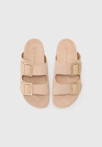 Beige semsket sklisikiner med to justerbare gullbelter, åpen tå, og polstret såle for komfort. Flat såle uten synlig mønster.