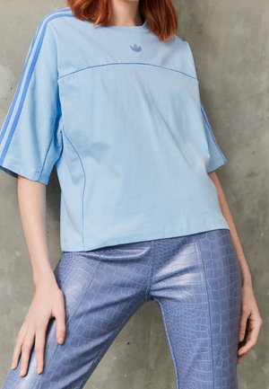 Rudowłosa osoba ubrana w jasnoniebieską koszulkę Adidas z paskami i logo, w połączeniu z błyszczącymi fioletowymi spodniami z imitacją skóry krokodyla, stoi przy szarej ścianie.