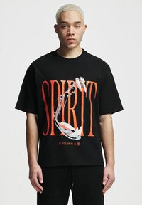 Juodas per didelis marškinėliai su ryškiu raudonu tekstu "SPIRIT", su spalvingu grafiniu dizainu ir logotipu apačioje. Trumpi rankoviai.