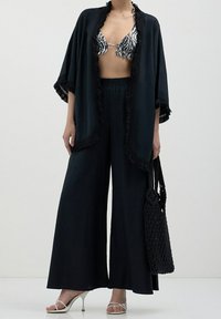 Czarny kimono z frędzlami, noszone na wzorzystym bikini i szerokich czarnych spodniach. Białe sandały na obcasie i czarna pleciona torebka dopełniają stylizację.