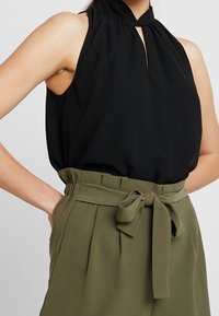 Top nero senza maniche con collo alto e dettaglio a chiave, abbinato a pantaloni verdi oliva a vita alta con vita annodata e frontale plissettato.