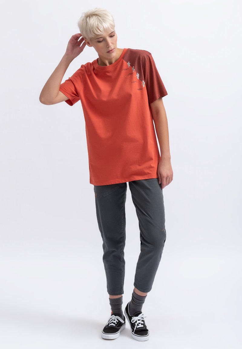 Een oranje en bruine t-shirt met korte mouwen en een grafisch ontwerp, gecombineerd met grijze tapered broek en zwarte sneakers. Soepele katoenstructuur.