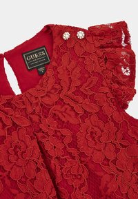 Rotes Spitzenoberteil mit floralen Mustern, Rüschensäumen und Perlenakzenten an den Knöpfen. Das Etikett trägt die Aufschrift "GUESS, EST. 1981, MODERNES DESIGN."