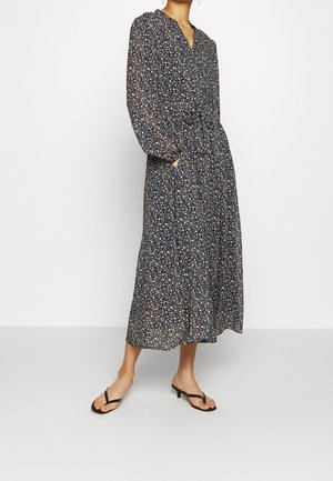 Kvinde iført en langærmet, ankel-lang blomsterkjole med taljeret pasform og sorte åbne hæle, stående på en ensfarvet hvid gulv.