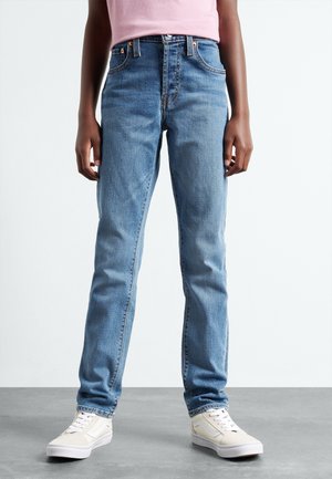 Person iført lyseblå jeans med lige ben, hvide sneakers og en lyserød top, stående på en grå gulv med hænderne afslappet langs siderne.