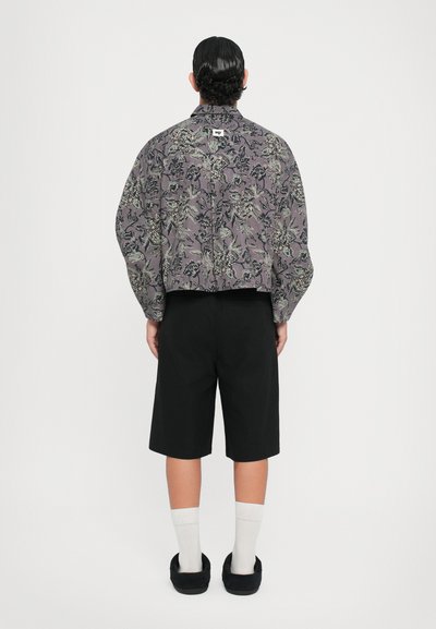 Blumenmuster, lila, übergroße Jacke mit Kragen, kombiniert mit schwarzen Shorts. Das Model trägt weiße Socken und schwarze Slipper.