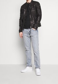 Giacca di pelle nera con colletto alto, abbinata a una t-shirt nera, jeans azzurri chiari e sneakers bianche. Texture liscia in tutta la composizione.