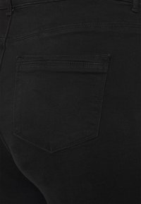 Jeans de mezclilla negra con un bolsillo trasero de diseño curvado y un ligero detalle de costura. La tela tiene una textura suave.