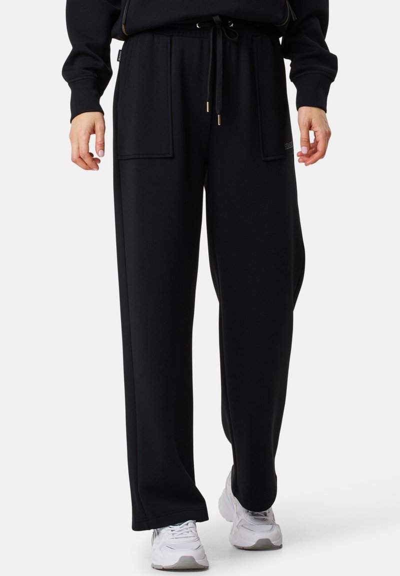 Sorte sweatpants med en afslappet pasform, der har to frontlommer, snoretræk i taljen og en glat stoftekstur. Båret sammen med hvide sneakers.