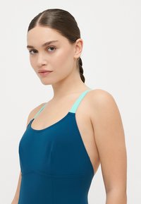 Fato de banho em tecido azul escuro com uma textura suave, apresentando alças em verde-água e um design desportivo. Vista aproximada da área do decote e dos ombros.