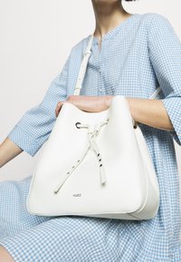 Bolso de cubo de cuero blanco con un acabado texturizado, correa ajustable, cierre de cordón y acentos plateados. Logotipo de la marca visible en la parte frontal.