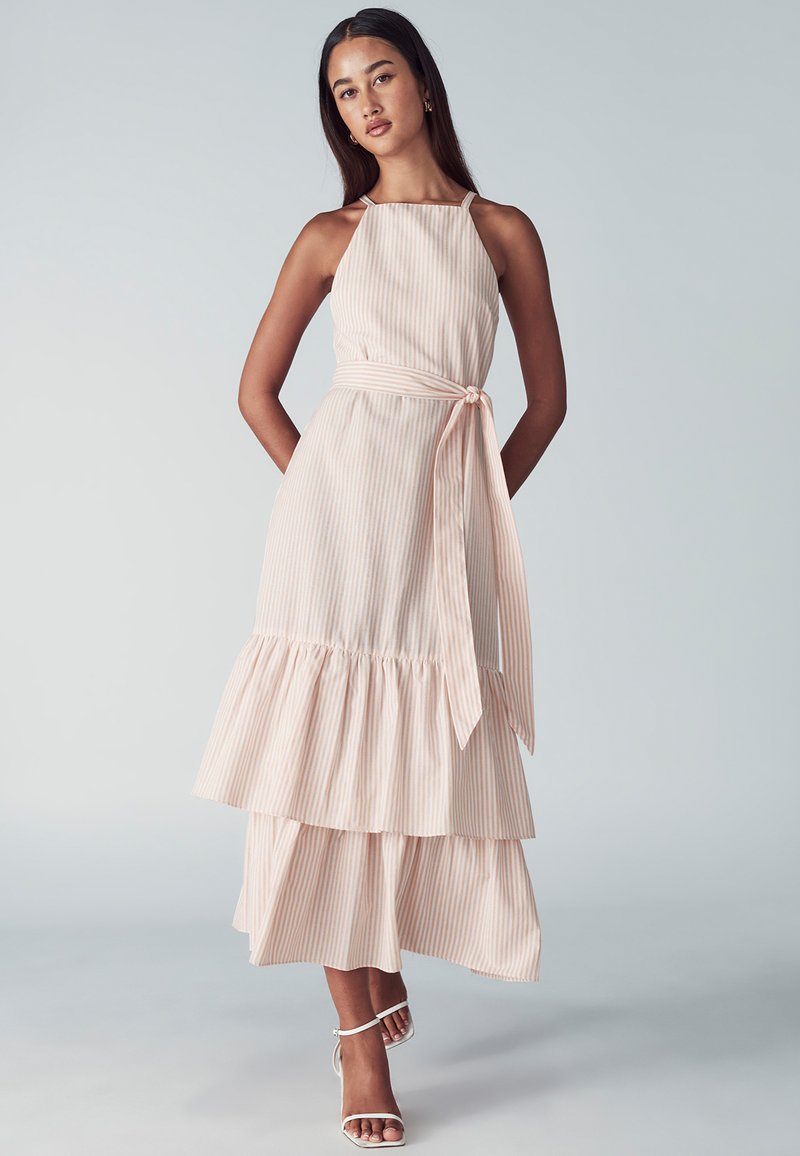Un vestido maxi a rayas de un rosa claro con cuello halter, dobladillo con volantes en capas y un detalle de cinturón atado en la cintura.