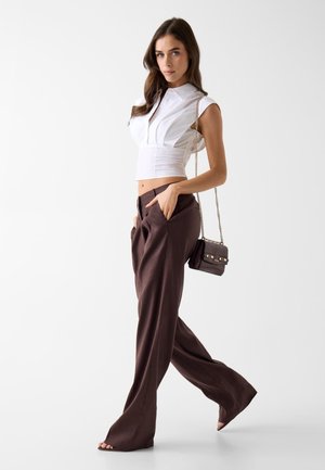 Donna che cammina indossando una camicetta bianca senza maniche e corta, e pantaloni marroni a gamba larga, con una piccola borsetta scura con tracolla a catena.