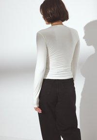 Bela dolgo rokava majica s prilegajočim krojem, oblečena z visokim pasom črnimi kavbojkami. Gladka tekstura, minimalni šivi. Senca, projicirana na steno.