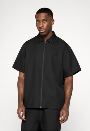 Mies, jolla on musta, lyhythihainen, vetoketjullinen kauluspaita, seisoo valkoista taustaa vasten.