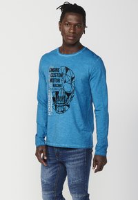Long-sleeve blue T-shirt in textured cotton, featuring a geometric skull graphic and text design in black and white on the front.