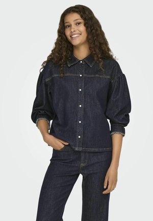 Chemise en denim bleu foncé avec un devant à boutons, des manches bouffantes et des coutures contrastées. Associée à un jean en denim assorti.