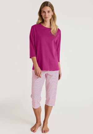 Vrouw staat blootsvoets en draagt een felroze top met lange mouwen en witte cropped broek met rode horizontale strepen tegen een effen achtergrond.