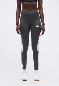 Γυναίκα που φοράει σκουρόγριες κολάν Adidas με λευκές ρίγες στο πλάι και λευκά αθλητικά παπούτσια, στέκεται μπροστά σε ένα απλό φωτεινό φόντο.