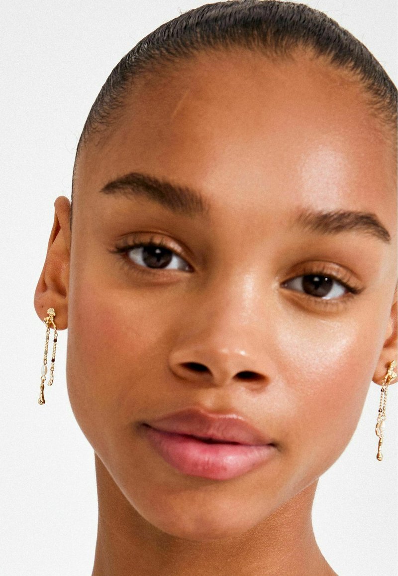 Gold drop earrings with a textured chain design and a small bead accent. Skin tone is medium with a smooth, radiant finish.