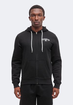 Jeune homme portant un sweat à capuche noir zippé avec des cordons blancs et un petit logo "Champion" sur la poitrine gauche.