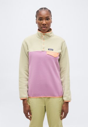 Žena v dvojfarebnom fleece pulóvri s béžovými, levanduľovými a broskyňovými detailmi, stojaca na jednoliatom bielom pozadí.