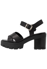 Sandales à talons en cuir noir avec deux brides croisées, une bride de cheville et une semelle épaisse texturée avec un léger compensé. Accent de boucle en argent.