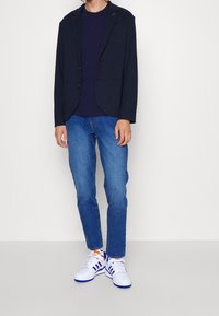 Marinblå kavaj med två framfickor, kombinerad med en mörk t-shirt, blå jeans och vita sneakers med blå och orange detaljer.