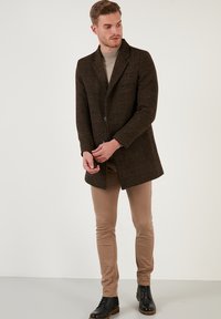 Abrigo de lana de cuadros marrón con cuello de muesca, cierre de un solo botón y bolsillos con solapa. Combinado con pantalones beige y botas negras con cordones.