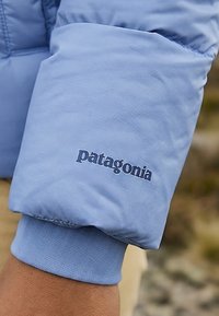 Ærme af lyseblå isoleret jakke med "patagonia" logo syet over en ribbet manchet på en persons arm.