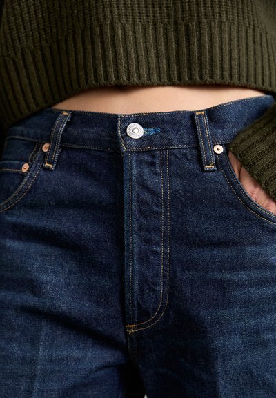 Dunkelblaue Jeans aus Denim mit taillierter Passform, silbernem Knopfverschluss und orangefarbenen Nähten. Kombiniert mit einem gerippten grünen Pullover.
