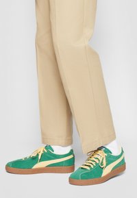 Baskets en daim vert avec des accents jaunes, des lacets texturés et des semelles en caoutchouc marron. Portées avec un pantalon beige et des chaussettes blanches.