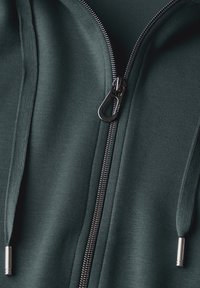 Hoodie zippé vert foncé avec une texture lisse, présentant une fermeture éclair en métal, des cordons de serrage et des accents argentés aux extrémités des cordons.