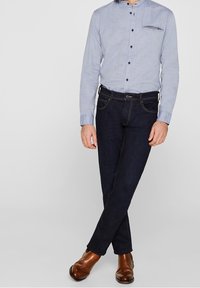 Camicia azzurro chiaro con colletto alla coreana, jeans blu scuro con cuciture a contrasto e scarpe in pelle marroni. Vestibilità slim e design moderno.