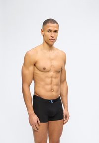 Νέος μυώδης άνδρας που φοράει μαύρα εσώρουχα Calvin Klein boxer, στέκεται μπροστά σε απλό φωτεινό φόντο, στραμμένος ελαφρώς προς τα δεξιά.