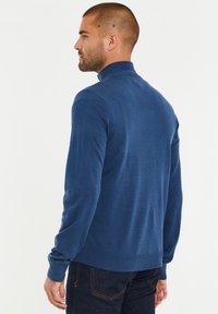 Blauer Strickpullover mit Stehkragen, strukturiertem Stoff, langen Ärmeln sowie gerippten Bündchen und Saum, kombiniert mit dunkelblauen Jeans.