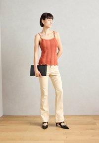 Top sans manches texturé couleur rouille avec un ourlet à volants, pantalon évasé beige, et sac à main et escarpins en cuir verni noir.