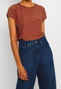 T-shirt à manches courtes de couleur rouille avec des motifs découpés au laser. Associé à un jean bleu foncé taille haute.