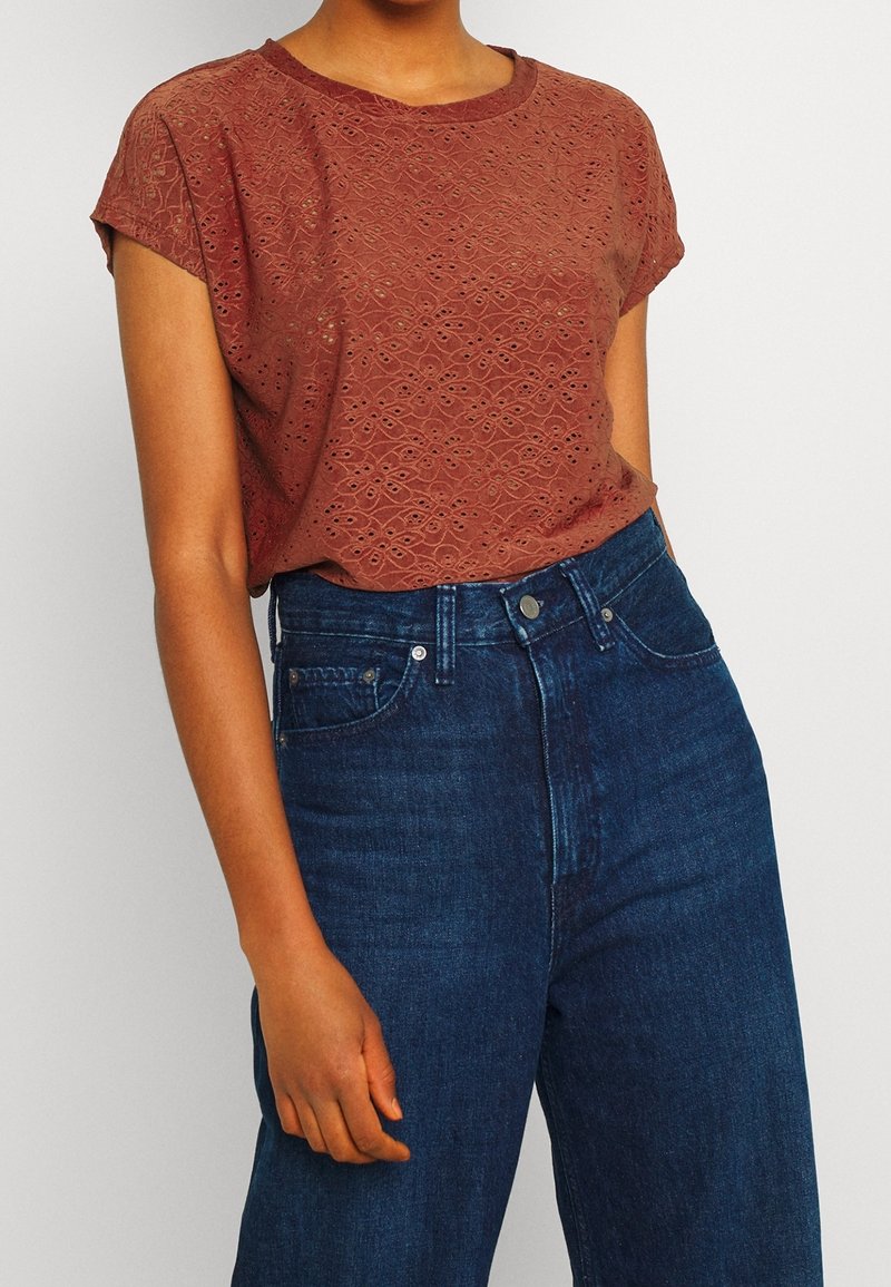 T-shirt à manches courtes de couleur rouille avec des motifs découpés au laser. Associé à un jean bleu foncé taille haute.